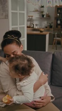 Sevgi dolu bir annenin, oturma odasındaki kanepede dinlenirken elmayı tutan ve onunla konuşan küçük kızı öptüğünün dikey görüntüsü.