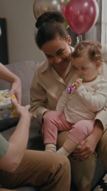 Babasının, annesinin kucağında, ağzında parti düdüğüyle oturan kızına doğum günü hediyesi verirken dikey bir fotoğraf.