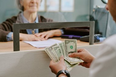 Banka şubesindeki cam bölmede müşterilerin mali işlemleriyle ilgilenen kıdemli bir banka memuru, yardım isterken para sayan bir müşteri.