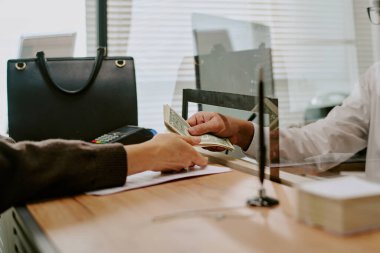 Tezgah üzerinden para değiş tokuşu yapan iki kişi, cam bölmeden para geçiyor, ödeme terminali ile ofis ayarlanıyor ve görülebilir belge yığını