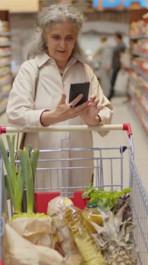 Gizemli alışverişçi olarak çalışan yaşlı bir kadının süpermarkette yürürken akıllı telefondan fotoğraf çekerken çekilmiş dikey görüntüleri.