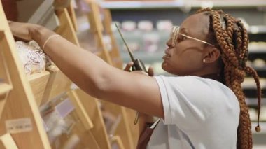 Süpermarkette çalışan genç siyahi kadının orta ölçekli yakın çekimi tezgahlardaki fırın ürünlerini kontrol etmesi ve telsizle iş arkadaşıyla konuşması.