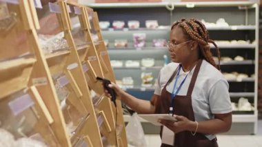 Süpermarkette çalışan genç Afrikalı Amerikalı dükkan asistanının tarayıcı ve dijital tablet kullanarak fiyat denetimi yaptığı orta boy bir fotoğraf.