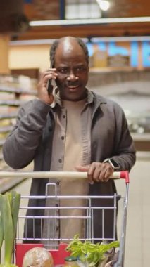Üst düzey siyahi adamın süpermarkette yemek yağı seçerken telefonda sohbet edişinin dikey görüntüsü.