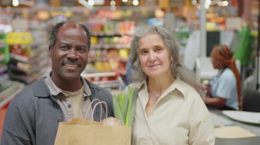 Markette kameraya gülümseyen ürünlerle dolu kağıt poşet taşıyan neşeli, etnik çeşitlilikte yaşlı bir çiftin portresi.