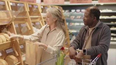 Neşeli son sınıf Kafkasyalı kadın ve Kara kocası modern süpermarkette yiyecek alışverişi yapıyorlar. Ekmek seçiyorlar.