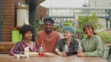 Çeşitli gen Z arkadaşlarının grup portresi sokak kafesinde yiyecek ve içeceklerle birlikte oturup, birlikte fotoğraf çektirip, yaz günü açık havada gülümsüyorlar.