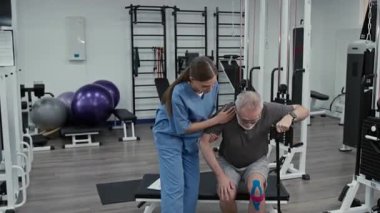 Genç bayan fitness koçu rehabilitasyon terapisi sırasında bastonlu yaşlı erkek müşterinin kürsüden kalkmasına yardım ediyor.