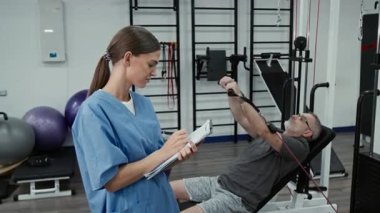 Olumlu beyaz kadın rehabilitasyon uzmanının portresi. Mavi önlük giyip kameraya gülümsüyor ve arka planda yetişkin hasta egzersizlerinin sonuçlarını yazıyor.