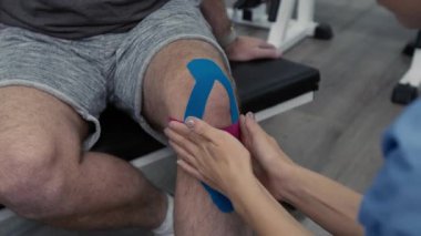 Rehabilitasyondan önce tanınmayan erkek hastaların diz kapaklarını bantlayan kimliği belirsiz genç kadın fizyoterapistin yakın çekimi.