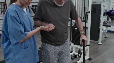 Genç bayan doktorun rehabilitasyon merkezinde bastonla yürürken iki eliyle yaralı erkek hastaya yardım edişini izleyin.