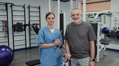 Milenyum kadın fizyoterapist portresi mavi önlüklü ve spor giyimli yaşlı erkek hasta rehabilitasyon merkezinde kameraya poz veriyor.