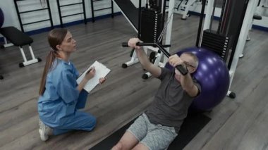 Olgun bir erkek hastanın kablo makinesinde öne doğru yükselişini gösteren yüksek açılı bir çekim. Mindere oturup egzersiz topuna yaslanırken, genç bayan fizyoterapist onu düzelterek ve sonuçları yazarken.