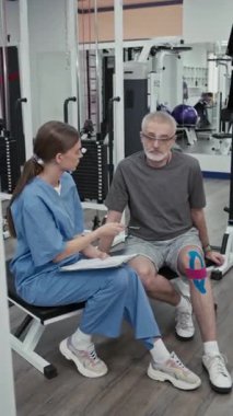 Mavi üniforma giyen genç bir bayanın dikey görüntüsü olgun bir adama diz yaralanması ve spor salonundaki rehabilitasyon egzersizinden önce yazma hakkında sorular sorması.