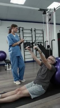 Olgun erkek hastayı kontrol eden kadın rehabilitasyon uzmanının dikey görüşü kablo makinesinde hafif ağırlıklarla omuz egzersizi yaparak eklemlerdeki sertliği azaltması.