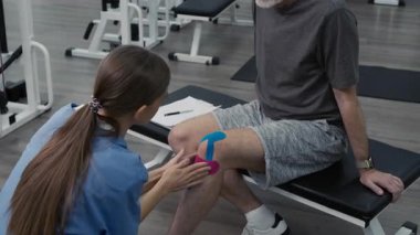 Uzun saçlı kadın fizyoterapist spor salonunda egzersiz masasında oturan olgun bir adamın dizine ağrı kesici bant takıyor.