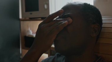 Yorucu bir iş gününden sonra mutfakta oturup gözlerini ovuşturan sinirli Afrikalı Amerikalı adamın yakın plan çekimi.