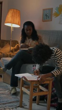 Dikey çekimde Afrikalı Amerikalı anne kitap okuyor ve oğluyla konuşuyor. Ödevini onunla kanepede yapıyor.