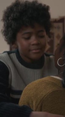 Afrikalı, kıvırcık saçlı, tanınmayan annesini kucaklayan ve kameraya bakan bir çocuğun dikey ağır çekim portresi.