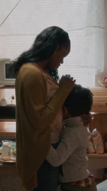 Afrika kökenli Amerikalı bir çocuğun annesine sarılırken, annesinin saçlarını okşadığı, mutfakta yan yana durduğu dikey fotoğraf.