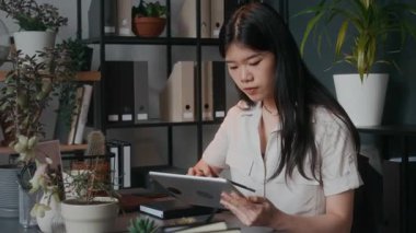 Arka planda bitki ve raflarla masa başında oturan genç bir kadın, iş günü rahat ofisinde dijital tablet kullanıyor.
