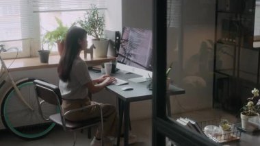 Modern ofisteki masaüstü bilgisayarında bitki ve bisikletle kod yazan genç bir kadın programcı.