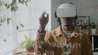 Ofiste VR kulaklık kullanan, sanal elementlerle etkileşim kuran, iş günü sanal gerçeklik uygulamasını test eden genç Afrikalı Amerikalı bir adamın cam duvarından bakın.