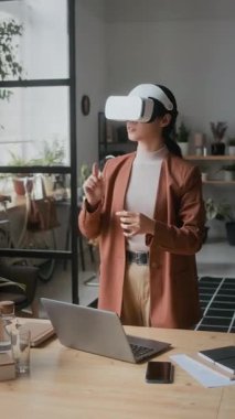 VR kulaklıklı kadın yazılım geliştiricisinin dikey görüntüsü bilgisayarla masada duruyor, sanal gerçeklikte çalışırken havada süzülüyor.
