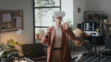 Bitkilerle süslenmiş modern hayvan dostu ofiste VR kulaklık kullanan genç bir kadın, arka planda çalışırken havadaki sanal elementlerle etkileşimde bulunuyor