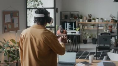 VR kulaklık takan genç siyahi bir adam modern ofiste sanal gerçeklik uygulamasını geliştirirken arka planda bilgisayar kodlaması yapıyor.