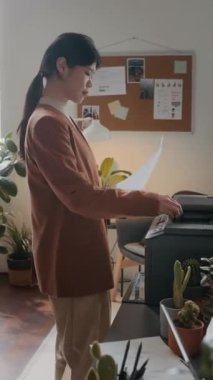 Asyalı genç bir ofis çalışanının, iç mekan bitkileriyle dekore edilmiş rahat çalışma alanındaki yazıcıya dokümanları kopyaladığı dikey bir fotoğraf.