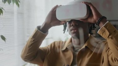 Modern ofiste VR kulaklığı takan genç siyahi bir adamın cam duvarından bakın. Büyük teknoloji şirketinde çalışırken artan gerçekliği deneyimliyor.
