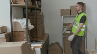 Yeşil yansıtıcı yelek giyen beyaz erkek çalışanı üç karton kutuyu alıyor ve iş günü boyunca onları depoda taşıyor.