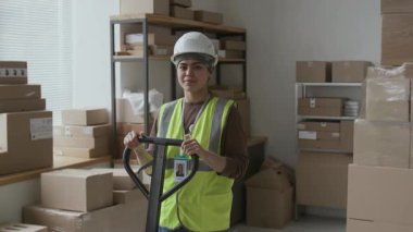 Yansıtıcı yelek giyen ve kameraya gülümseyen kasklı İspanyol kadın işçi portresi depoda elle kaldırma aletini kullanıyor.