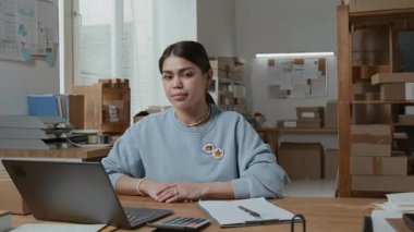 Z jenerasyonu kadın depo çalışanının portresi mavi kazaklı, kameraya gülümseyen rozetliler üzerinde dizüstü bilgisayar, hesap makinesi ve pano olan ahşap masada oturuyor.