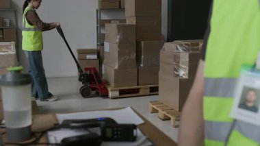 Odaklan, depoda çalışan bilinmeyen bir erkek arkadaş bilgisayarla uğraşırken ağır karton kutularla manüel kaldıraç kullanan bir kızın görüntüsünü yakınlaştır