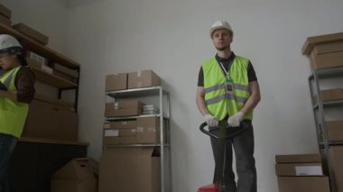 Düşük açılı erkek çalışanın yüksek görüşlü yelek giymesi ve kalın kasketli ağır yükleri el paleti kamyonuyla taşıması ve kadın çalışana iş günü depoda hızlıca kafa sallaması.