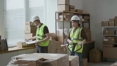 Karton kutularla manuel forklift taşıyan kadın işçi, erkek meslektaşı ona gelip depodaki sevkiyat belgelerini kontrol ediyor.