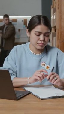 Genç bayan çalışanların dizüstü bilgisayardaki işlemci emirlerine dikey bir bakış açısı ve depo masasındaki bir kağıt parçasına veri yazma