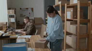 İspanyol Gen Z kızı karton kutuyu bantlıyor ve depoda çalışırken ahşap rafa koyuyor. Beyaz erkek meslektaşı arka planda dizüstü bilgisayarla yazı yazıyor.