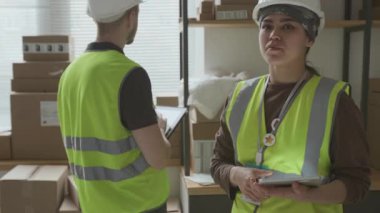 Dijital tabletli, yeşil güvenlik yeleği giyen ve beyaz şapkalı bir kız erkek arkadaşının yanından geçiyor ve depodaki büyük kutuda barkod taraması yapıyor.