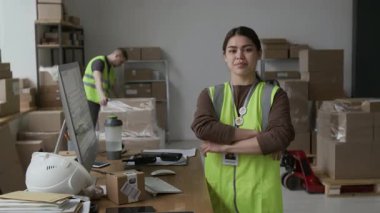 Pozitif İspanyol asıllı genç bir kadının portresi. Yeşil, yüksek görünürlüklü, kollarını kavuşturmuş ve iş günü depoda kameraya bakıyor.