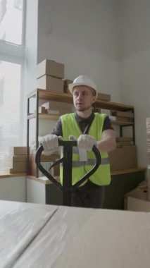 Beyaz şapka ve yeşil güvenlik yeleği giyen beyaz erkek yükleyicinin dikey görüntüsü depoda kutularla el paleti kamyonu taşıyor.