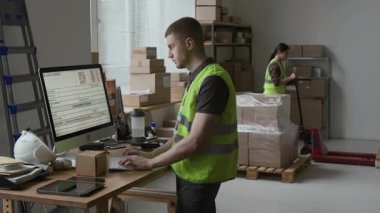 Yeşil yansıtıcı yelek giyen erkek işçi küçük bir paket alıyor ve depoda bilgisayar üzerinde çalışırken numarasını giriyor, arka planda elle kaldırma kullanıyor.