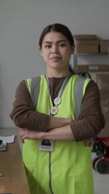 Yansıtıcı yelek giyip iş yerinde kameraya gülümseyen neşeli esmer depo müdürünün dikey portresi.