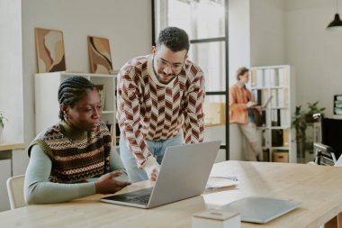 Orta yaşlı, yakışıklı erkek müteahhitin, Afrikalı Amerikalı bayan meslektaşına dizüstü bilgisayarında şifreli olarak yardım ederken çekilmiş bir fotoğrafı.