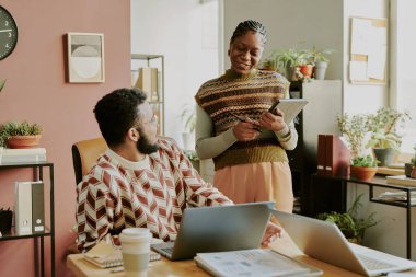 Orta boy, elinde tablet olan Afro-Amerikalı bir kadının masada dizüstü bilgisayarla çalışan bir adama baktığı bir fotoğraf.