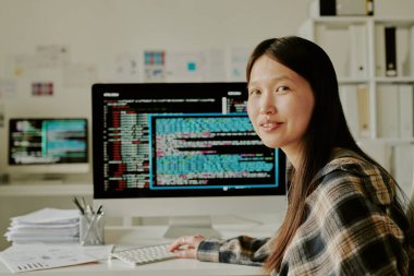 Çekici gülümseyen genç programcının portresi çalışma masasında oturmuş arka planda kod satırlarını gösteren dev bir monitörle.