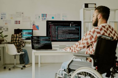 Ofiste kodlama yaparken dev ekranlara bakan engelli programcının orta boy görüntüsü.