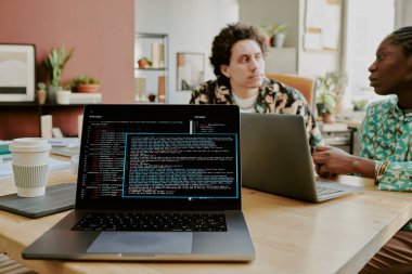 Ekranda kod çizgileri olan gümüş dizüstü bilgisayarı kapat. Arka planda iki melez bilişim bürosu çalışanı konuşuyor.